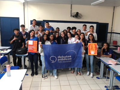 Grupo de jovens reunidos em uma sala de aula. A turma sorri para a foto e mostra a bandeira dos Debates Públicos nas Escolas