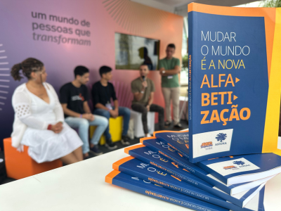 Em foco, à direita, há vários exemplares do livro "Mudar o Mundo é a Nova Alfabetização". Ao fundo, há cinco jovens sentados em pufes conversando