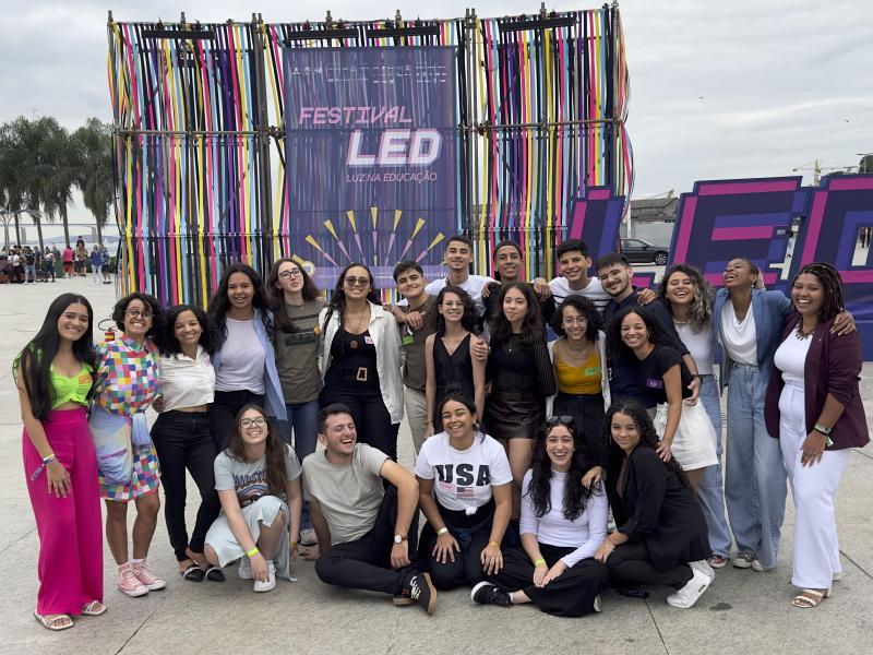 Grupo diverso de jovens reunidos ao ar livre. Atrás do grupo, há uma estrutura metálica colorida